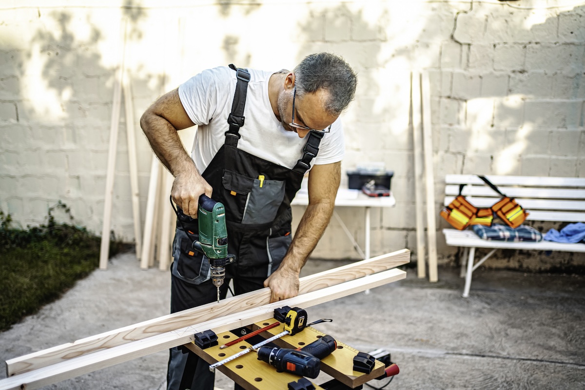 Mature male carpenter using electric drill on a wooden plank out